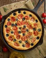 Focaccia, pizza in skillet, italian flat bread with tomatoes, olives and rosemary. Wooden table