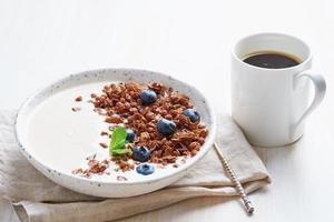 Yogurt with chocolate granola, bilberry. Breakfast with cupof coffee on a white background, side view