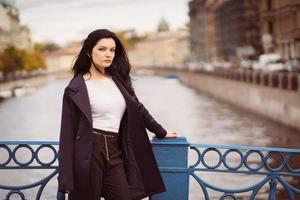Charming thoughtful fashionably dressed woman with long dark hair travels through Europe, standing in city center of St. Petersburg. A beautiful girl wanders alone through autumn streets photo