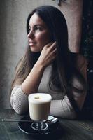 Beautiful girl sits in cafe in Christmas holidays, in background lights of garland. Brunette woman with long hair drinks cappuccino coffee, latte and looks out window, dark backdrop photo