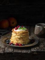 Pancake with vanilla cream, blueberries and raspberries. Side view, vertical, copy space. Dark moody old rustic wooden background.