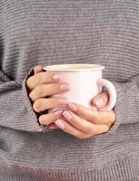 café caliente de la mañana en el trabajo en una mañana fría de otoño, manos sosteniendo una taza con una bebida, suéter gris, manicura rosa, primer plano, vertical