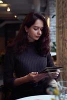 Beautiful serious stylish fashionable smart girl sit in cafe in loft style on lunch. Ready to place an order, studying menu. Charming thoughtful woman with long dark brown hair. photo