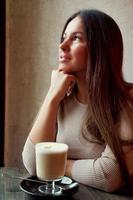 Beautiful pensive happy girl sitting in cafe in Christmas holidays, smiling and dreaming. Brunette woman with long hair drinks cappuccino coffee, latte and looks out window photo