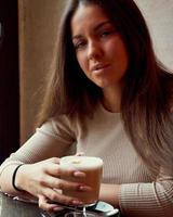 Beautiful pensive happy girl sitting in cafe in Christmas holidays, smiling and dreaming. Brunette woman with long hair drinks cappuccino coffee, latte and looks to camera