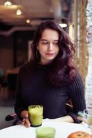 hermosa chica seria, elegante y elegante está sentada en un café y bebiendo un saludable batido verde amarillo o vegano con leche. encantadora mujer pensativa con largo cabello castaño oscuro. foto