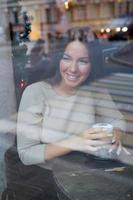 A beautiful girl sits in a cafe and looks out window thoughtfully. Reflection of city in window. Smiling brunette woman with long hair drinks cappuccino coffee, vertical photo