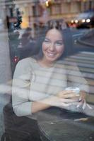 una hermosa chica se sienta en un café y mira por la ventana con cuidado. reflejo de la ciudad en la ventana. mujer morena sonriente con el pelo largo bebe café capuchino, vertical foto