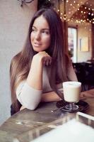 Beautiful girl sits in cafe in Christmas holidays, in background lights of garland. Brunette woman with long hair drinks cappuccino coffee, latte and looks out window, dark backdrop