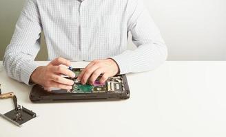 el hombre repara la computadora. un ingeniero de servicio con camisa repara una computadora portátil, en un escritorio blanco contra una pared blanca. foto