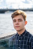 Portrait of young guy, brunette with dark hair, Caucasian. Close - up of a guy, boy, man looking at camera. Outdoor in city by water photo