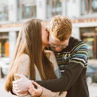 Portrait of happy couple embracing in downtown, red-haired man with glasses kisses and woman with long hair. Girl whispers in ear of guy. Concept of teenage love