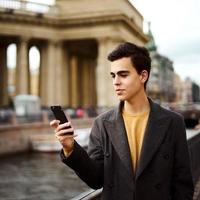 Handsome stylish fashionable man talking on phone, dialing chat message, brunette in elegant gray coat is standing on street in historical center. Young man with dark hair, thick eyebrows. photo