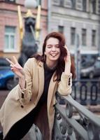 Beautiful serious stylish fashionable smart girl standing on bridge and smiling, show tongue, show victory sign with hand. St. Petersburg city. Charming funny happy woman with long dark hair photo