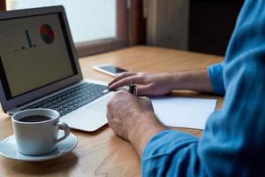 Unrecognizable man writes plan on paper and looks at charts on computer screen, laptop. A man with casual clothes in blue shirt sits in office in front of window photo