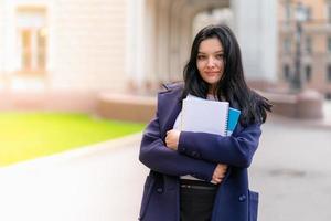 Beautiful smiling girl brunette student holding notebooks and textbooks, stands at University on street of St. Petersburg. A charming woman with long dark hair is studying at course photo