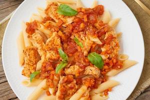 Penne pasta, chicken or turkey fillet, tomato sauce with basil leaves on old rustic wooden background. Top view, close up photo