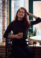 hermosa chica seria, elegante y a la moda se sienta junto a la ventana en el café, sonríe maravillosamente y bebe batido vegano saludable o café con leche. encantadora mujer pensativa con largo cabello castaño oscuro foto