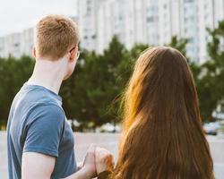 Girl with long thick dark hair holding hands redhead boy in blue t-shirt on bridge, teen love at evening. Boy looks tenderly at girl, young couple. Concept of teenage love and first kiss