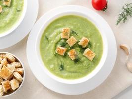 Two large white bowl with vegetable green cream soup of broccoli, zucchini, green peas on white background, top view photo