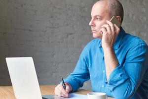 Handsome handsome mature man looks out window and talking on cell phone, sitting at computer, laptop. Man with casual clothes in blue shirt and table at office in front of window, copy space photo