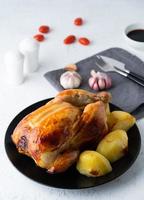 Grilled whole chicken in black plate on white table, baked meat with potatoes. Side view, vertical photo