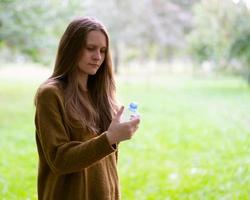 hermosa joven bebiendo agua de una botella de plástico en la calle en el parque en otoño o invierno. una mujer con hermoso cabello largo y oscuro mira una botella de agua
