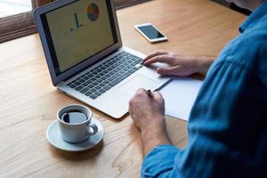 Unrecognizable man writes plan on paper and looks at charts on computer photo