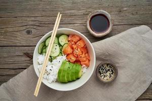 Salmon poke bowl with fresh fish, rice, cucumber, avocado with black and white sesame. Old wooden table. Food concept. photo