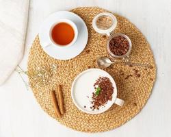 Yogurt with chocolate granola in cup, breakfast with tea on beige background, top view.