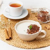 Yogurt with chocolate granola in cup, breakfast with tea on beige background, side view