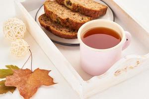 comida de otoño: rebanadas de pan de plátano, una taza de té, hojas secas, mesa de madera blanca. acogedor hogar de invierno. vista lateral, espacio de copia