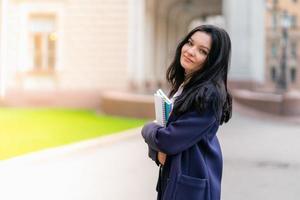 Beautiful smiling girl brunette student holding notebooks and textbooks, stands at University on street of St. Petersburg. A charming woman with long dark hair is studying at course, copy space photo