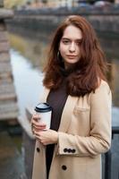 Beautiful serious stylish fashionable girl holding cup of coffee in hands walking street of St. Petersburg in city center. Charming thoughtful woman with long dark hair looks at camera photo
