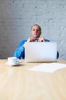 Handsome handsome mature man sitting at table in front of laptop and listening to subordinate. Man with casual clothes in blue shirt in the office in front of window, vertical photo