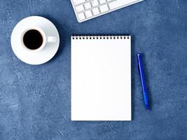 el bloc de notas abierto con una página blanca limpia, un bolígrafo y una taza de café sobre una mesa de piedra azul oscuro envejecida, vista superior