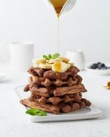 gofres de chocolate y plátano con flujo de jarabe de arce en jarra de leche, crema en mesa blanca foto