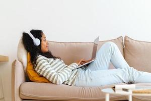 African-American female in headphones surfing the net on laptop while lying on sofa in living room photo