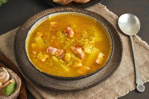 Winter hot soup with chopped green peas, pork, bacon, smoked on dark brown wooden table. Delicious appetizing fat rich soup.
