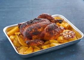 Baked whole chicken in blue casserole on dark blue table, roasted meat with potatoes. Side view photo