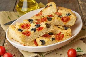 Focaccia, pizza in plate with tomatoes, olives and rosemary. Chopped Italian flat bread.