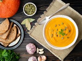 pumpkin soup mashed in a white plate, pumpkin seeds, bread, garlic on dark wooden background photo