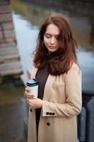 Beautiful serious stylish fashionable girl holding cup of coffee in hands walking street of St. Petersburg in city center. Charming thoughtful woman with long dark hair looks down, vertical photo