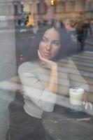 A beautiful girl sits in cafe and looks out window thoughtfully. Reflection of city in window. Brunette woman with long hair drinks cappuccino coffee, vertical photo