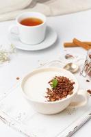 yogur con granola de chocolate en taza, desayuno con té sobre fondo blanco de madera, vertical. foto