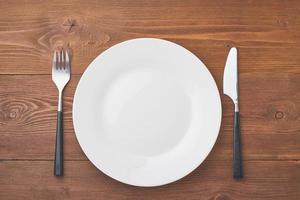 clean empty white plate, fork and knife on brown wooden table, copy space, mock up photo