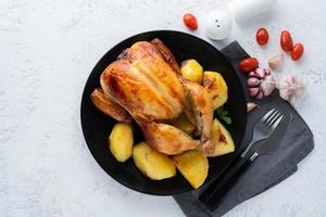 pollo entero a la parrilla en plato negro sobre mesa blanca, carne al horno con patatas. vista superior, espacio de copia foto
