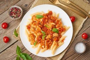 Penne pasta, chicken or turkey fillet, tomato sauce with basil leaves on old rustic wooden background. Top view photo
