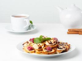 Belgian curd waffles with raspberries, banana, chocolate syrup. Breakfast with tea on white background, side view photo