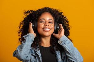 People, music, emotions concept. Delighted carefree female with Afro hairstyle dances in rhythm of melody, closes eyes listens loud song in headphones. Beautiful afro woman with her headphones photo